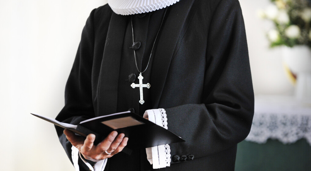 Nærbillede af præsts torso. På brystet bærer præsten et kors og i hænderne er der en bønnebog