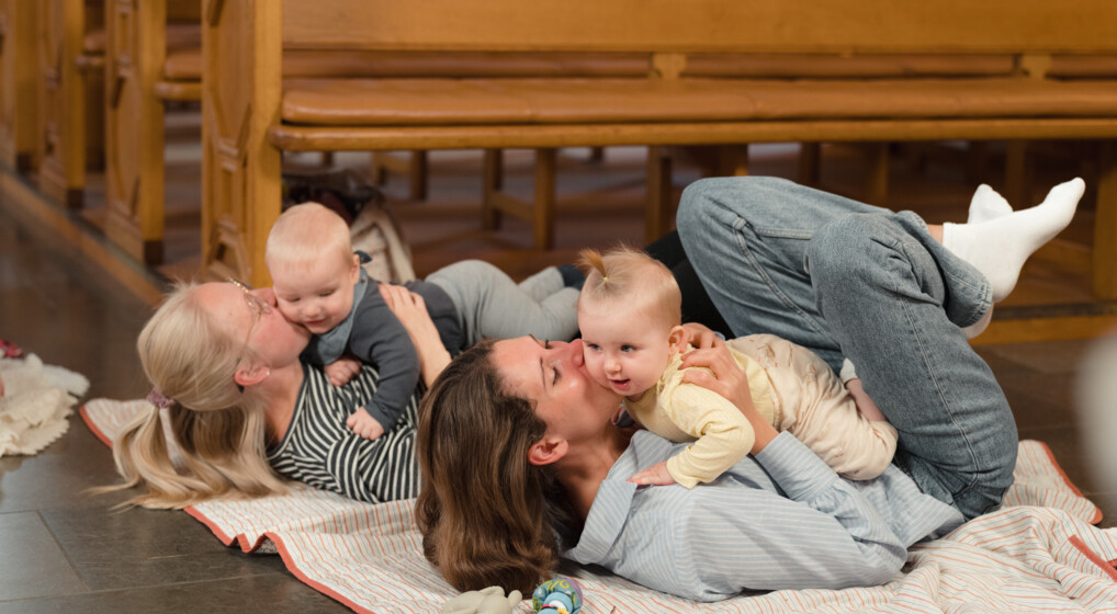 To mødre ligger på ryggen på gulvet med deres babyer på maven til babysalmesang i en kirke