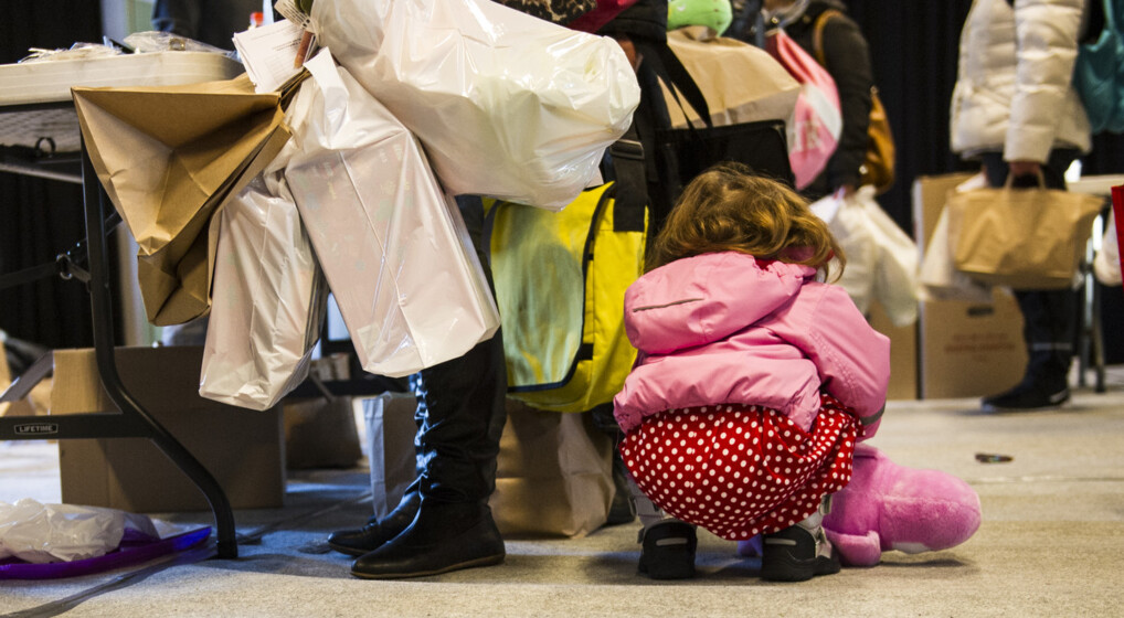 Barn bøjer sig ned for at samle sin bamse op ved siden af en kvinde med favnen fuld af indkøbsposer