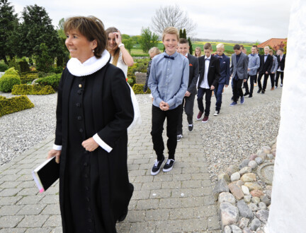 En præst går foran en lang række glade konfirmander uden for en kirke.