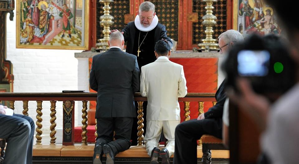 To mænd iklædt jakkesæt knæler foran alteret, hvor biskop Kjeld Holm er ved at vie dem