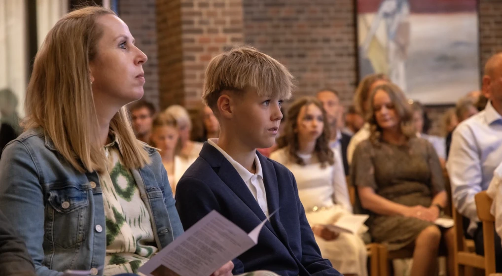 En konfirmand og hans mor sidder i kirken og lytter opmærksomt under en konfirmation.