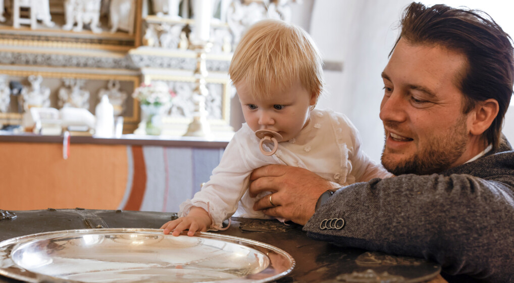 Far med dåbsbarn i dåbskjole ved dåbefonten