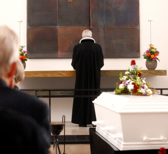 En præst står ved alteret under en bisættelse i kirken. I forgrunden ses en hvid, blomsterpyntet kiste.