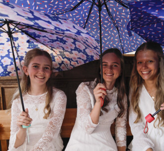 Tre smilende konfirmander sidder under to farvestrålende paraplyer.