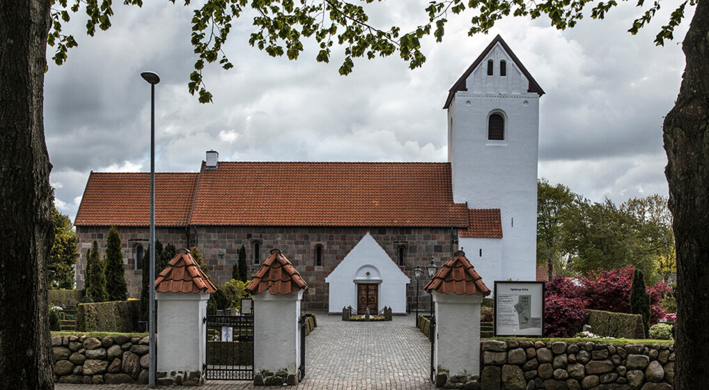 Middelalderkirke bygget af kvadre med hvidkalket tårnomgivet af kirkegård og træer.